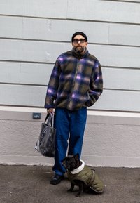 Fleecová bunda s károvaným vzorom v tmavozelených, fialových a béžových farbách, skombinovaná s modrými džínsami a čiernymi topánkami; sprevádzaná malým psom v zelenom kabáte.