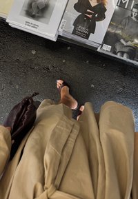 Rochie trench bej, până la genunchi, cu talie legată, purtată cu sandale slide violet închis. Într-o mână se ține o geantă pouch violet închis.