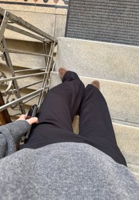 Black trousers, grey patterned jumper, brown shoes, and a dark phone held against grey stone steps and a metal railing.