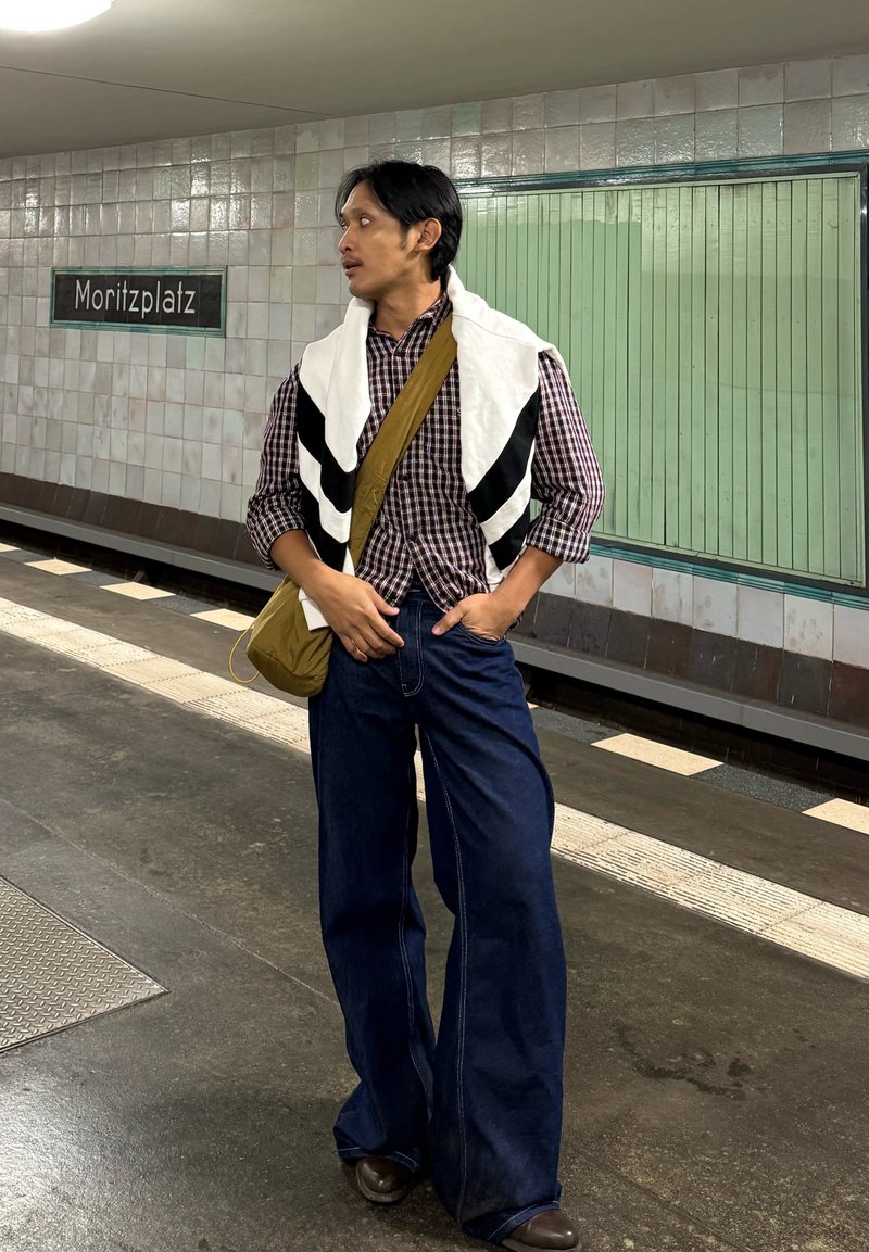 Homme portant une chemise à carreaux, un pantalon en jean surdimensionné et une serviette blanche à rayures noires posée sur ses épaules. Sac messager marron.
