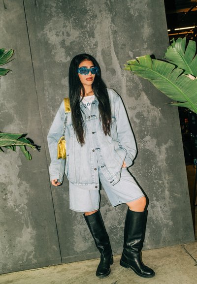 Chaqueta de mezclilla azul claro sobre un conjunto de dos piezas a juego, combinada con botas de cuero negras hasta la rodilla y gafas de sol azul oversized.