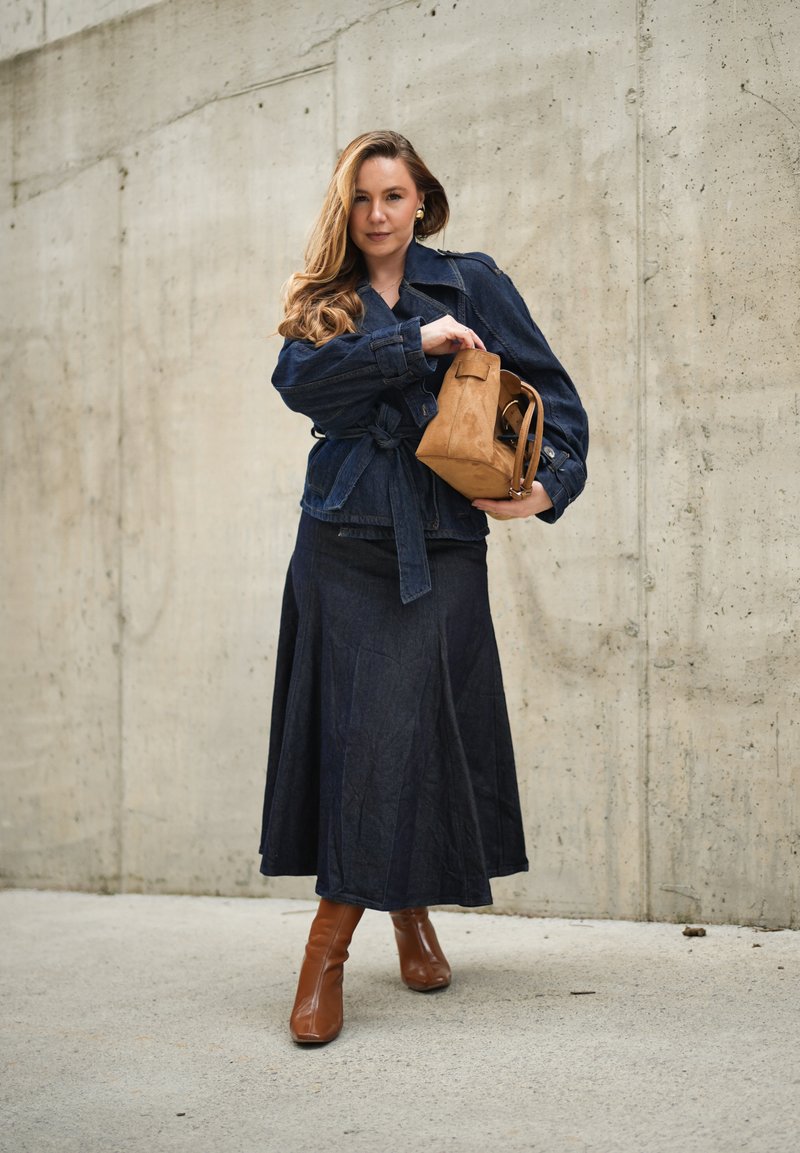 Femme en veste en jean foncée et jupe tenant un sac à main en daim beige, portant des bottines marron, debout devant un mur en béton.