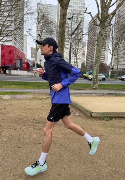 Hombre corriendo en un parque de la ciudad, con gorra negra, chaqueta azul, pantalones cortos negros, calcetines blancos y zapatillas de correr verde claro. Edificios altos al fondo.