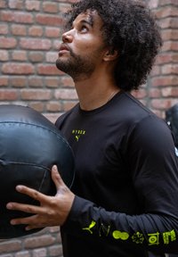 Must musta pika varrukaga särk helerohelise logoga, hoides musta meditsiinipalli, taustal tekstuuriga sein punaste tellistega.