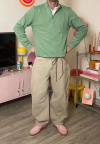 Svetlozelený sveter na zips, béžové nohavice so šnúrkou a ružové topánky. Pozadie zdobí farebná výzdoba a nábytok.