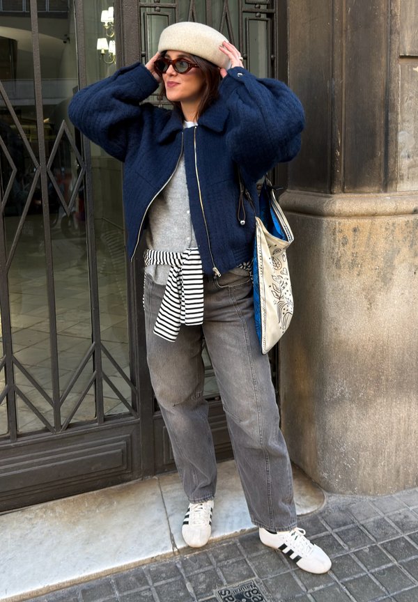 Vrouw met zonnebril, beige baret, marineblauwe donsjas, grijze spijkerbroek, gestreept shirt dat om de taille is geknoopt, witte sneakers en een draagtas, die haar hoed buiten rechtzet.