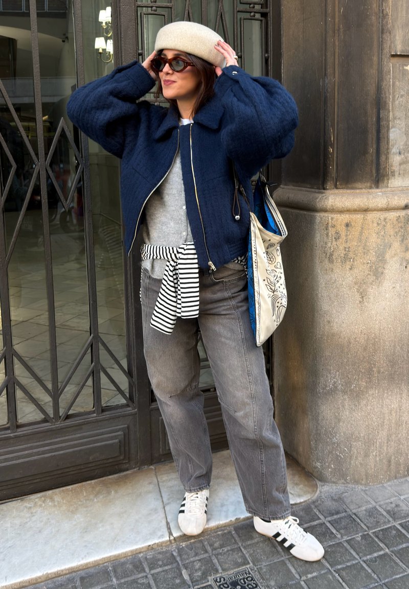 Femme portant des lunettes de soleil, un béret beige, une doudoune bleu marine, un jean gris, une chemise rayée nouée à la taille, des baskets blanches et un sac tote, ajustant son chapeau en plein air.