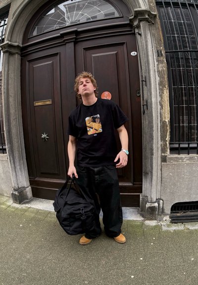 Camiseta negra con un diseño gráfico en la parte frontal, combinada con pantalones negros de corte suelto y zapatos marrones. Bolso de deporte negro grande sostenido en la mano.