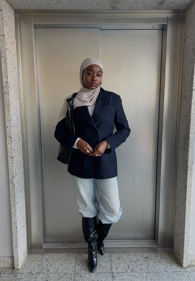 Blazer a rayas marino sobre una blusa blanca, combinado con pantalones cortos de mezclilla claros y botas altas negras, complementado con un bolso negro.