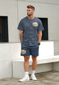 Maglietta a maniche corte grigia in rete e pantaloni corti con il logo "Los Angeles Lakers" in giallo e blu. Indossati con scarpe da ginnastica bianche e calzini.