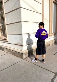Camicia a maniche lunghe viola con il logo "Lakers", pantaloni corti in denim neri e sneakers grigie con dettagli rossi. In piedi all'angolo di un edificio beige.