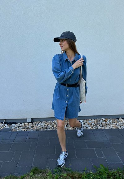 Vestido camisa de mezclilla con mangas largas, botón en la parte delantera y cintura ceñida con cinturón. Combinado con una gorra negra y zapatillas deportivas, llevando una bolsa de tela.