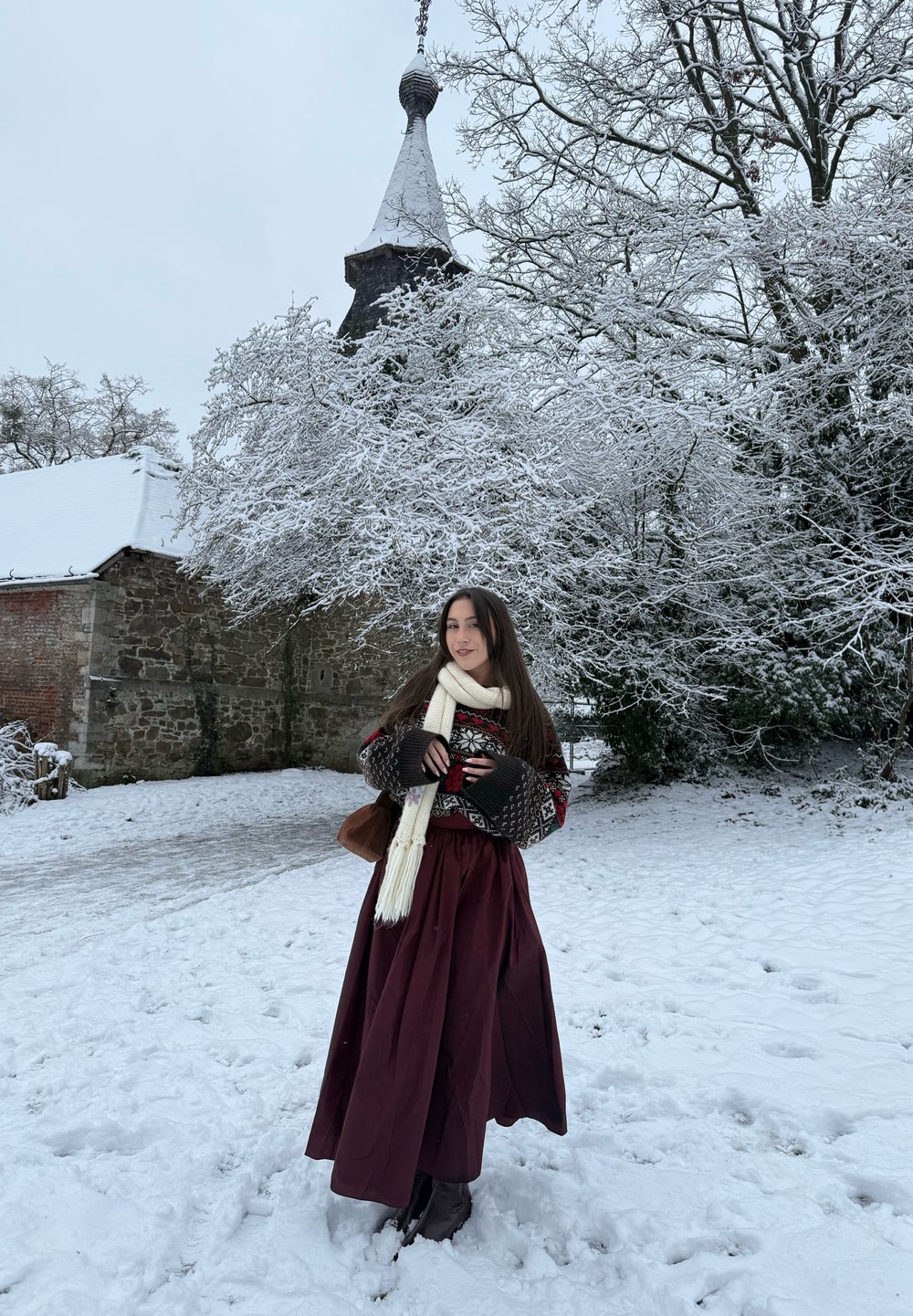 Ung kvinde iført en mønstret sweater, hvidt tørklæde og lang bordeaux nederdel står på snebelagt jord nær en stenmuret bygning og snefyldte træer.