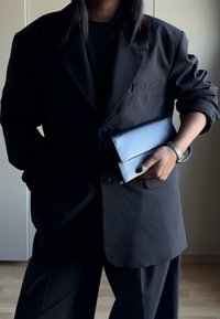 Blazer noir à coupe décontractée porté sur un haut noir, tenant une pochette bleue pliée. Textures remarquables : tissu lisse et ferrures polies.