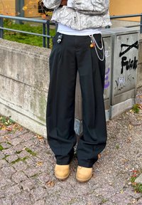 Čierne široké nohavice s hladkou textúrou, zdobené kovovými reťazcami s malými príveskami. Kombinované s hnedými dreváčkami na textúrovanom povrchu.