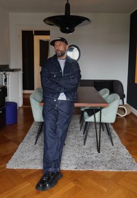Een man staat met gekruiste armen in een kamer, gekleed in een donkerdenim jasje en bijpassende broek, met zwarte schoenen, op een gestructureerd grijs vloerkleed.
