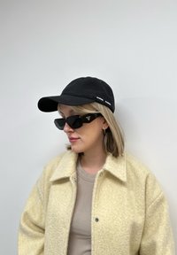 Μαύρο καπέλο μπέιζμπολ με λευκό κείμενο, χοντρή μπεζ fleece ζακέτα, ανοιχτό καφέ ριγωτό μπλουζάκι και γωνιακά μαύρα γυαλιά ηλίου με λεπτομέρεια λογότυπου.