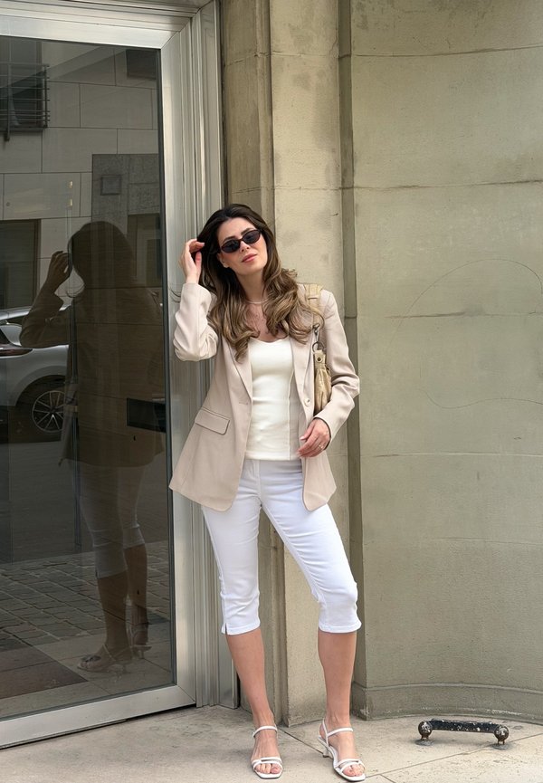 Vrouw met zonnebril, beige blazer, wit topje en capri-broek, poserend tegen een beige muur bij een glazen deur in een stedelijke omgeving.