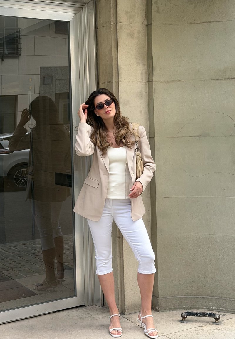 Frau mit Sonnenbrille, beigem Blazer, weißem Top und Caprihose, die vor einer beigen Wand in der Nähe einer Glastür in urbaner Umgebung posiert.