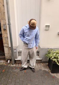 Šviesiai mėlyni marškiniai ilgomis rankovėmis su sagų uždarymu, pilkos laisvos kelnės ir ruda kepurė su juoda užrašu „EAST“. Rudi batai.