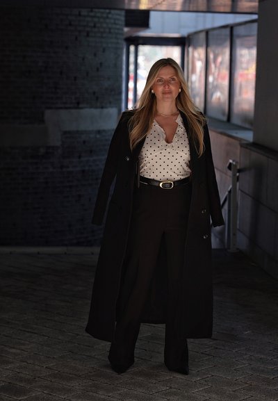 Mujer con cabello rubio largo de pie en un túnel oscuro, vestida con abrigo negro, blusa blanca con lunares, pantalones negros y pendientes de perlas.