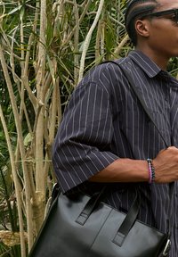 Borsa a spalla nera in pelle con manici piatti, indossata sopra una camicia a righe navy a maniche corte. Braccialetto di perline colorate sul polso.