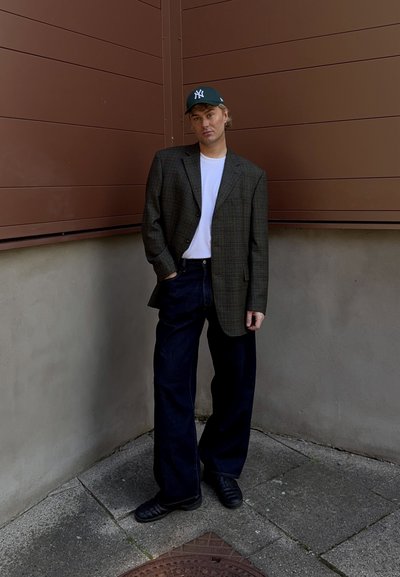 Blazer de cuadros, camiseta blanca, jeans de pierna ancha oscuros y zapatos negros. Lleva una gorra verde con un logotipo blanco. De pie contra una pared marrón.