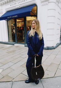 Ένα σκουρό μπλε ραμμένο κοστούμι με φαρδιά παντελόνια, μακρύ παλτό και καφέ τσάντα στον ώμο. Το φόντο απεικονίζει ένα κατάστημα με σκούρο μπλε τέντα.