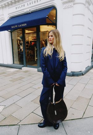 Un traje azul oscuro a medida con pantalones anchos, un abrigo largo y un bolso marrón de hombro. El fondo muestra una tienda con un toldo azul marino.