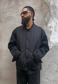 Schwarze gepolsterte Weste mit Knopfverschluss, Seitentaschen und glatter Textur, über einem schwarzen Shirt getragen, vor einem strukturierten grauen Hintergrund.