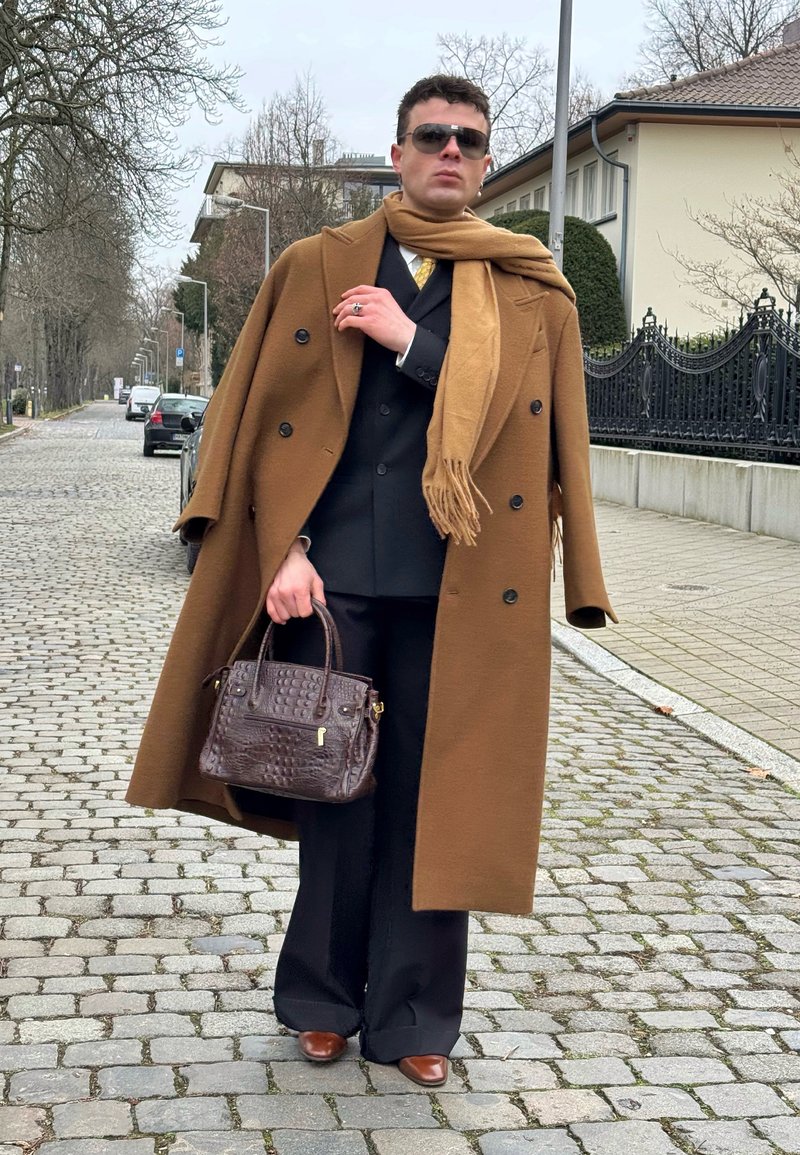 Man loopt op een kasseienstraat, draagt een bruine overjas en sjaal, een zwart pak, zonnebril en houdt een donkerbruine leren handtas vast.
