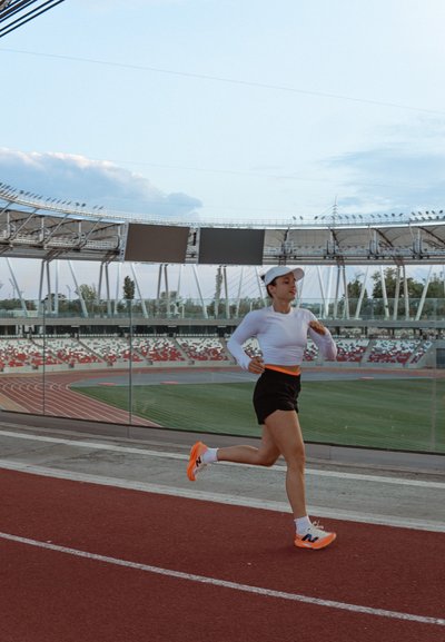 Atleta con una camiseta de manga larga blanca, pantalones cortos negros y zapatillas con detalles en naranja, corriendo en una pista roja con un fondo de estadio.
