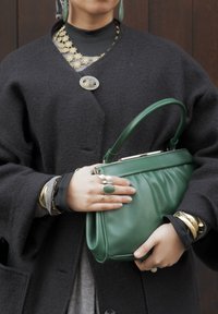 Personne portant un manteau sombre et des bijoux en superposition tenant un sac à main en cuir vert avec une poignée courte devant un fond en bois.