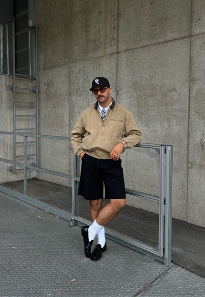 Chaqueta bomber beige, camisa blanca, corbata estampada, pantalones cortos negros, zapatos negros y calcetines blancos; de pie contra una pared de hormigón con barandilla de metal.
