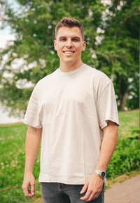 Μπεζ βαμβακερό T-shirt με κοντά μανίκια, με εκτύπωση κειμένου στο μπροστινό μέρος. Φορεμένο με σκούρα τζιν και ασημένια ρολόι χειρός. Πράσινο φόντο.