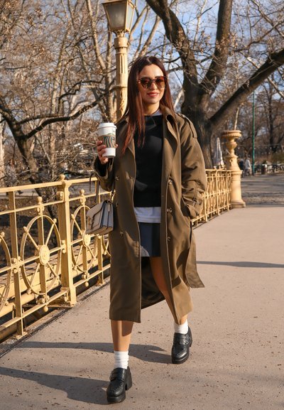 Gabardina verde oliva sobre un suéter negro, falda gris y calcetines blancos. Zapatos de plataforma negros. Sujetando una taza de café. Camino de piedra y farolas.