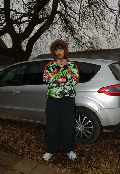 Persona vistiendo una sudadera gráfica verde con diseños de calaveras, pantalones negros y zapatillas plateadas, de pie junto a un coche plateado.