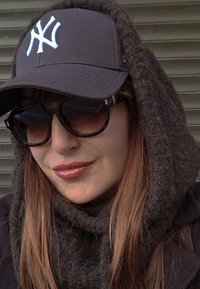 Fekete baseballsapka fehér logóval, nagy méretű sötét napszemüveg és barna kötött sál. Simaságot sugárzó textúra és meleg színvilág.