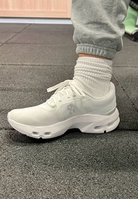 Fehér atlétikai cipők, texturált hálós felsőrésszel, lekerekített orral, vastag talppal és finom márkajelzéssel. Fehér bordázott zoknival és szürke nadrággal viselve.