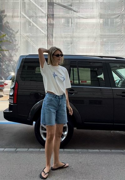 Camiseta blanca de manga corta con diseño de estrella azul, combinada con pantalones cortos de mezclilla azul claro y chanclas negras, al lado de un vehículo negro.