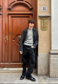 Jean noir et pull gris sous un manteau foncé, debout à côté de portes en bois. Un interphone en laiton est fixé sur le mur à côté d'eux.