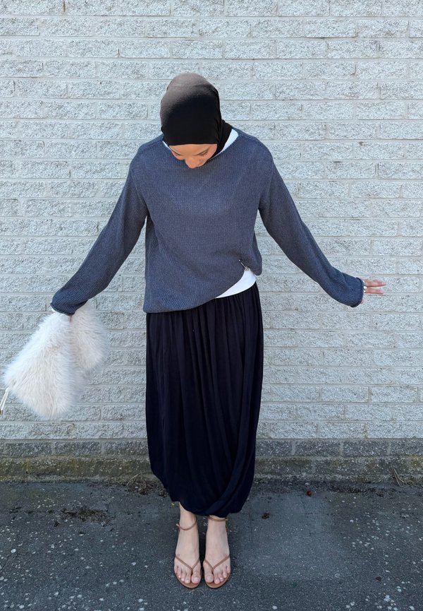 Femme portant un foulard sombre, un pull gris ample, une longue jupe noire et des sandales, se tient devant un mur en briques gris clair en tenant un sac blanc et duveteux.