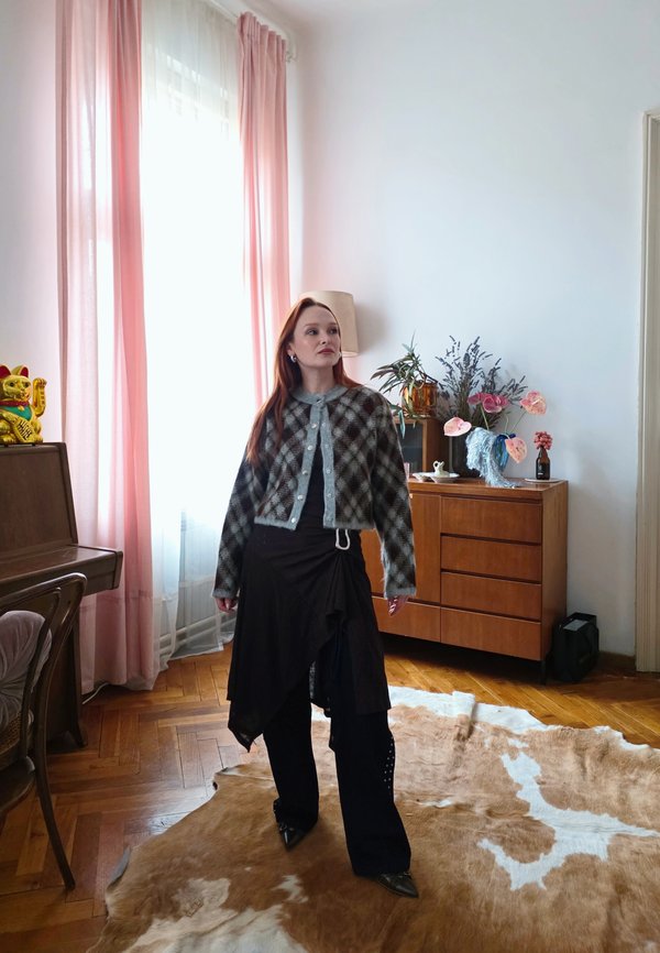 Sieviete ar sarkaniem matiem, tērpusies rūtainā kardiganā un slāņainā melnā svārku komplektā, stāv uz govs ādas paklāja telpā ar rozā aizkariem un koka mēbelēm.