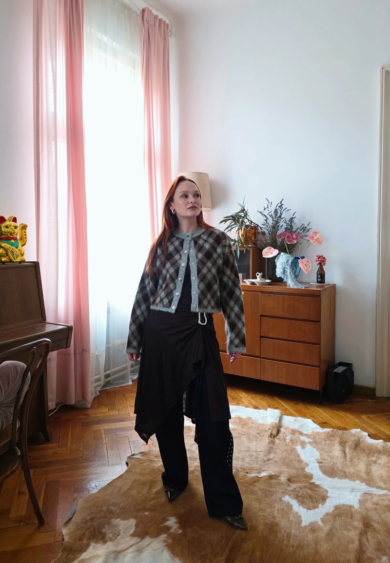 Femme aux cheveux roux portant un cardigan à carreaux et une jupe noire en couches, debout sur un tapis en cuir de vache dans une pièce avec des rideaux roses et des meubles en bois.