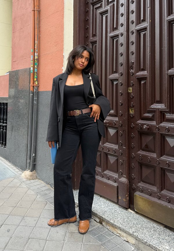 Femme en tenue noire et chaussures brunes appuyée contre une grande porte en bois ornée sur un trottoir urbain tenant un gobelet bleu.
