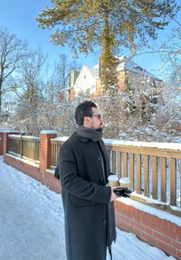 Homme portant un manteau noir et une écharpe, tenant deux tasses de café et des gants, debout sur un chemin enneigé à côté d'une clôture en briques, avec des arbres recouverts de neige et une maison en arrière-plan.