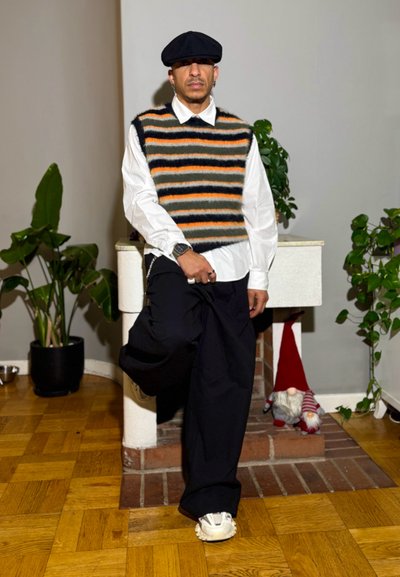 Chaleco de punto con rayas horizontales en gris, naranja y negro sobre una camisa blanca; combinado con pantalones negros de pierna ancha y zapatillas blancas.