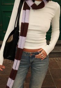 Kobieta ubrana w białą bluzkę z długim rękawem, niebieskie dżinsy, w paski brązowy i jasnofioletowy szalik, srebrne pierścionki oraz czarną torebkę na ramię, stojąca na zewnątrz.