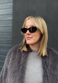 Sorte katteøyne solbriller med en blank finish, kombinert med en myk grå faux pelsjakke og en lys grå genser. Glatte, teksturerte stoffer.