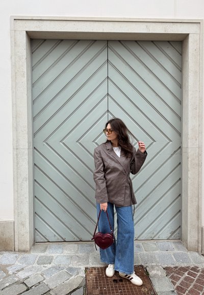 Blazer cruzado marrón, camisa blanca, vaqueros azul ancho, bolso burdeos en forma de corazón, zapatillas blancas, de pie frente a una puerta chevron verde claro.